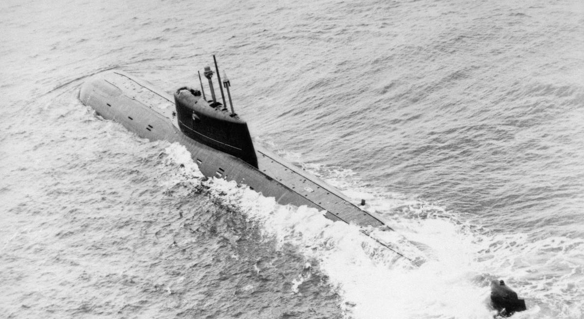 Чёрно-белый снимок с самолёта: длинная подводная лодка идёт в надводном положении, над корпусом высокая рубка с выдвинутыми мачтами, за кормой пенный след
